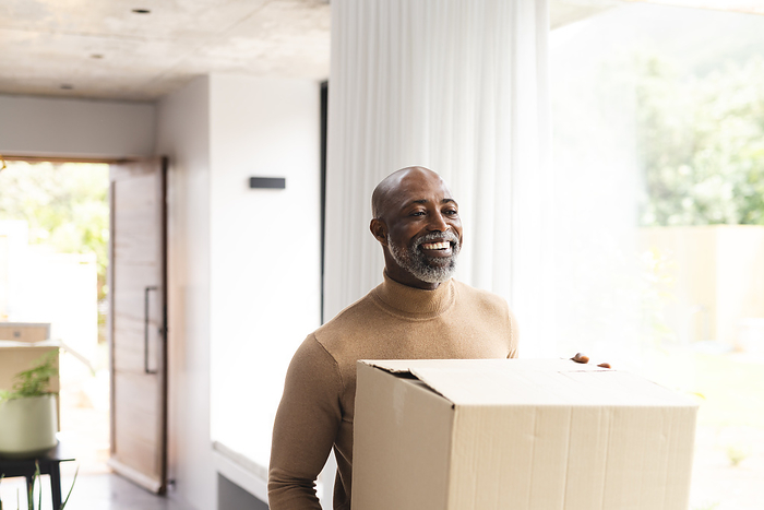 Happy african american mature man carrying packing box into new home, copy space. Home ownership, property, moving house, domestic life and lifestyle, unaltered.
