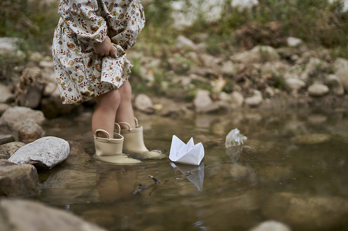 Girl wearing rubber boots and playing with paper boat in water puddle
