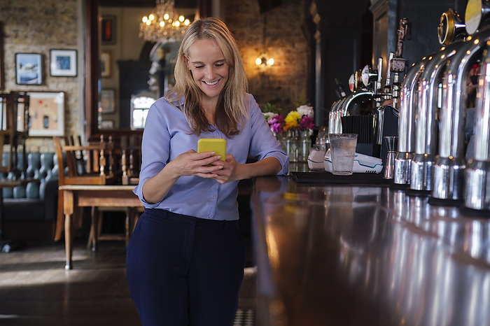 Smiling owner using smart phone near bar counter at pub