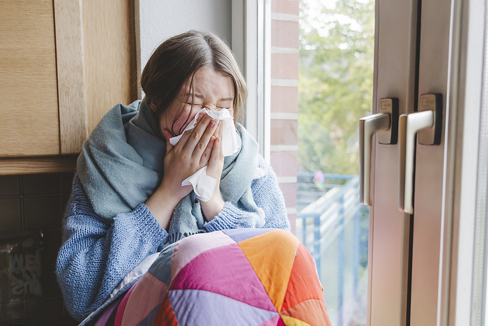 Sick girl blowing nose on tissue near window at home