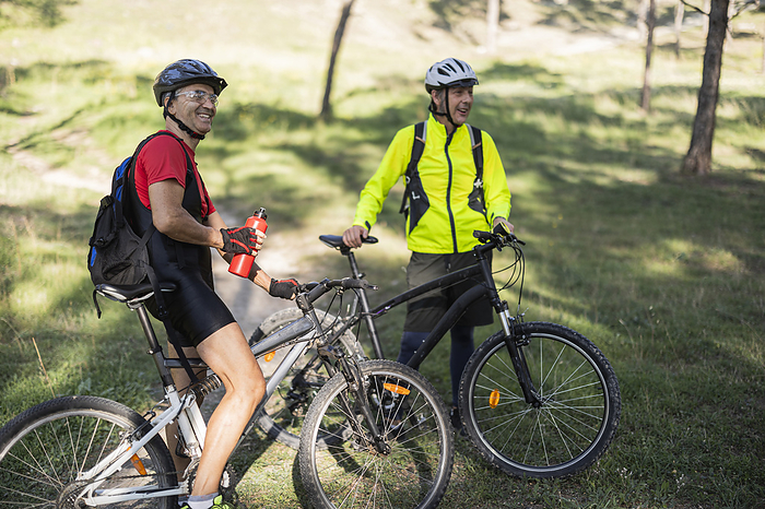 Smiling man holding water bottle and standing near friend with bicycle in forest