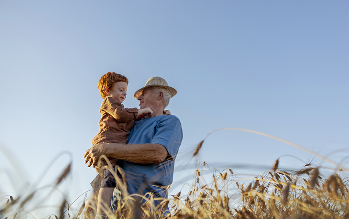 Grandfather wearing straw hat and carrying grandson under sky