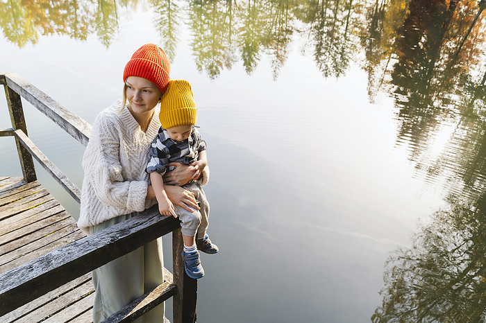Smiling mother holding son sitting on wooden railing near lake