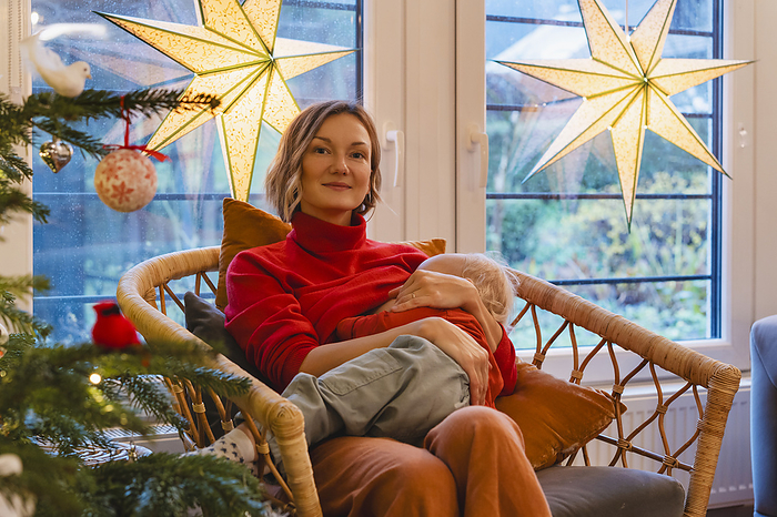 Blond woman with sleeping son sitting on chair at home during Christmas