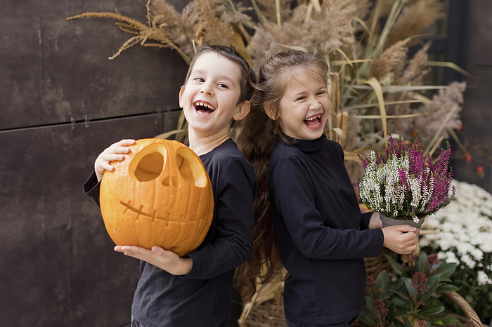 Cheerful siblings holding carved pumpkin and flower pot in hand near wall
