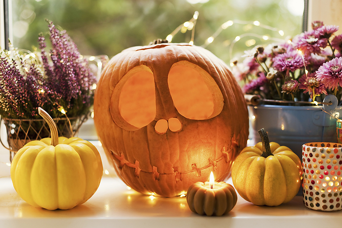 Halloween decoration of Jack O' Lantern with pumpkins and flowers near candle on window sill
