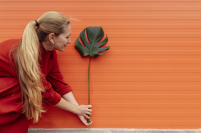 Blond woman holding monstera leaf and crouching in front of orange wall