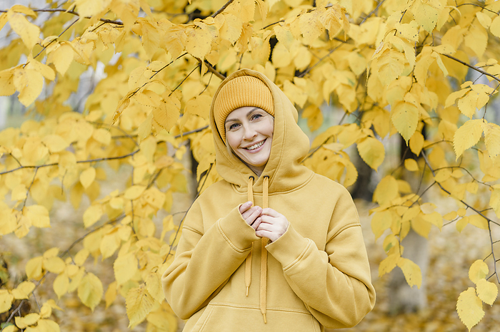Happy woman wearing hooded shirt and knit hat in front yellow leaves at park