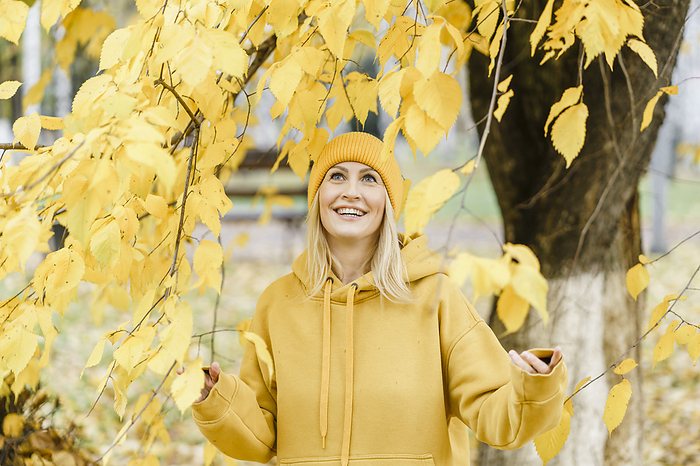 Smiling woman wearing yellow hooded shirt and touching leaves of tree in park