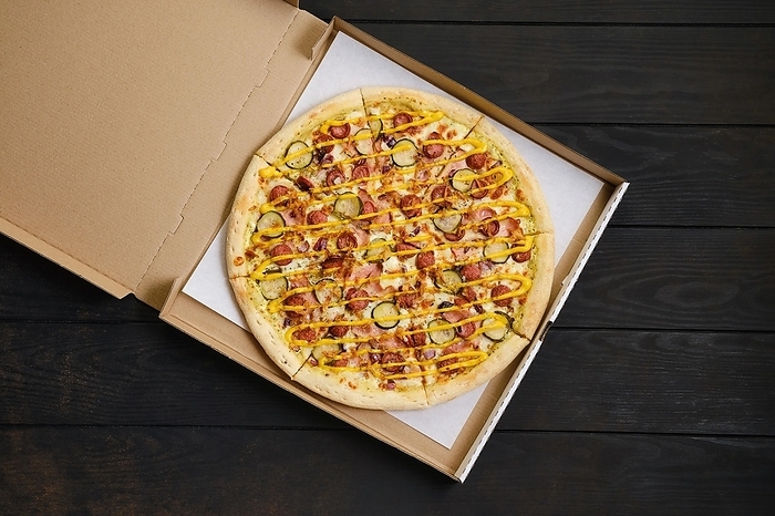 Top view of pizza with sausage, ham, pickled cucumber, onion and mustard in cardboard package