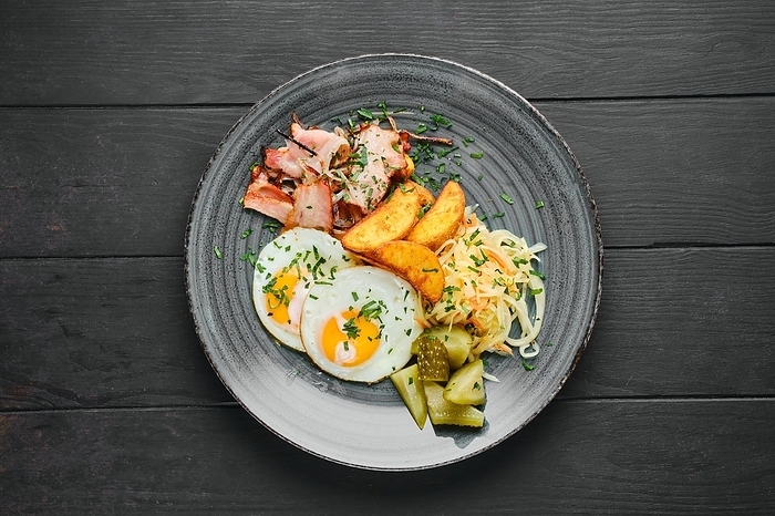 Overhead view of fried eggs, ham with potato wedges, pickled cabbage and cucumber on black wooden table