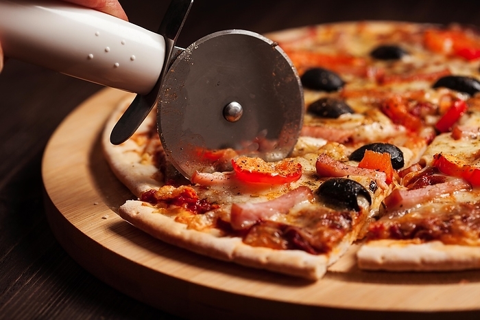 Pizza cutter wheel slicing ham pizza with (capsicum) and olives on wooden board on table
