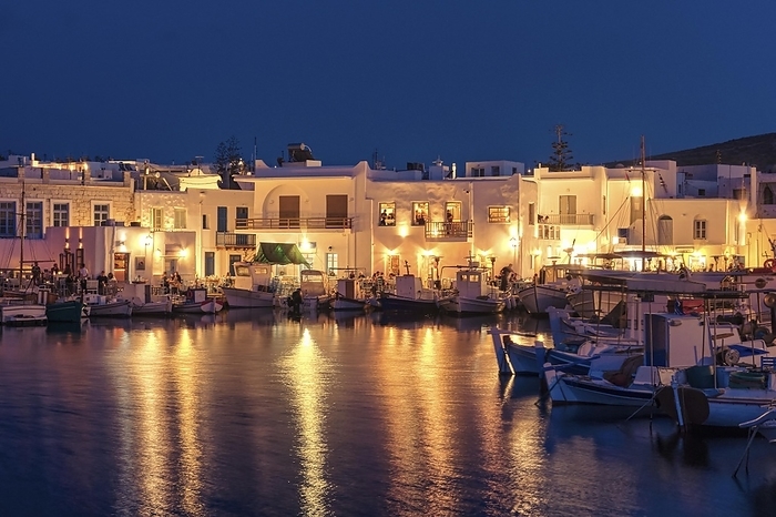 Traditional Greek fishing village and harbour after sunset, boats moored by jetty, streetlights and whitewashed houses alongside. Naoussa, Paros island, Greece. Clear blue sky, colorful reflections, summer vacation vibe