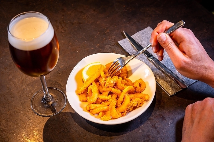 A customer eating squid breaded with lemon accompanied with a beer on a black background, on a white plate, by Unai Huizi