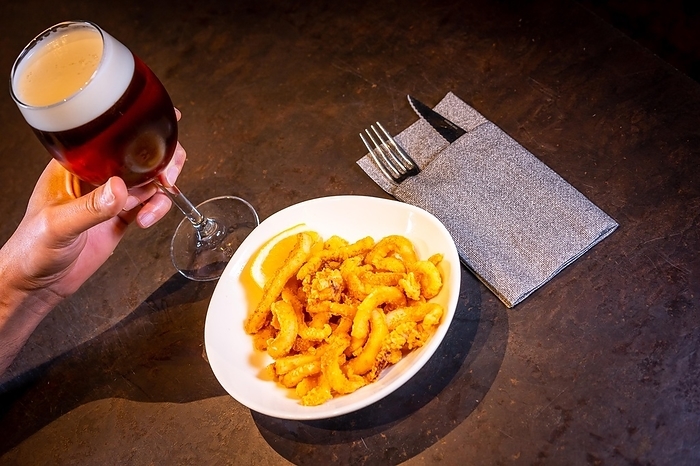 A customer eating squid breaded with lemon accompanied with a beer on a black background, on a white plate, by Unai Huizi