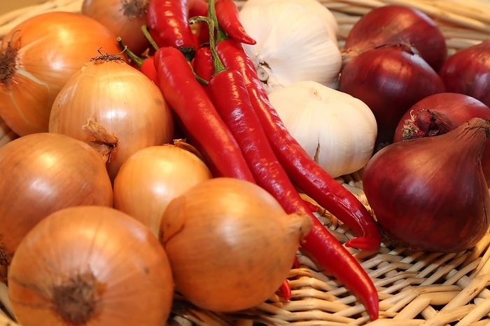 A woven basket filled with red chilli peppers, white garlic and yellow onions. The basket is made of woven straw. The chilli peppers are long and slender with a pointed tip. The garlic is large and spherical. The onions are medium sized and round, by fotoping