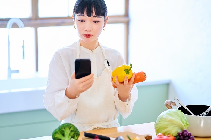 Young Japanese woman holding vegetables and smartphone in her kitchen (People)