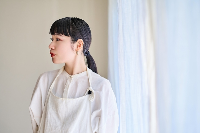 A young Japanese woman in an apron resting by the window of her room.