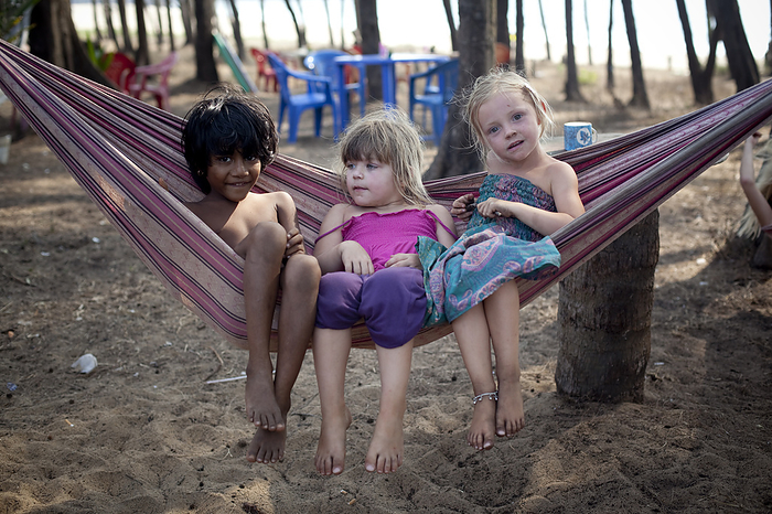 India British Kids on holiday play in a hammock and make friends with a local girl on Turtle Beach, Goa, India., by Vicki Couchman   Design Pics