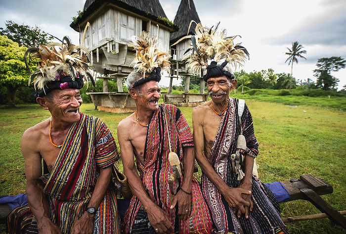 Group Of Men In Traditional Attire Share A Laugh With Sacred Houses In The Background; Lospalmos District, Timor-Leste, by David Kirkland / Design Pics