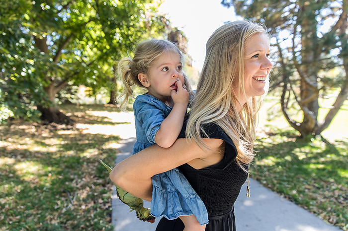 Mom smiling giving daughter piggy back ride on sidewalk at park, by Cavan Images / Rachel Greiman
