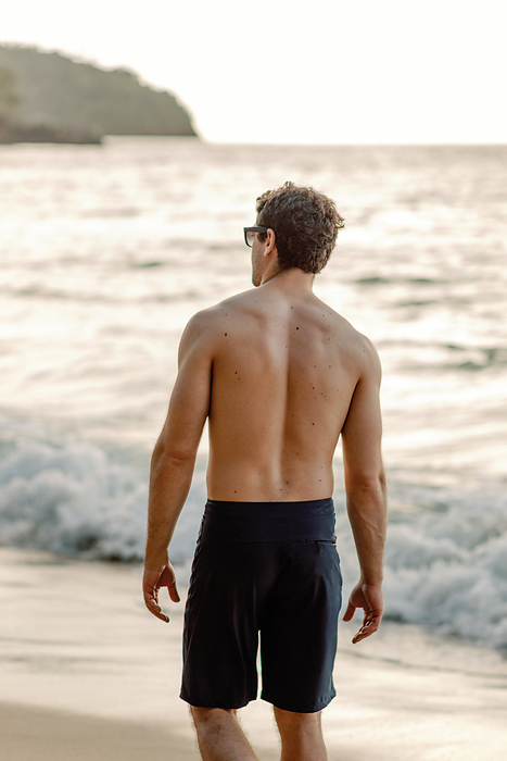 Muscular back, man in swimsuit walks Dominican beach at sunset., by Cavan Images / Samantha Joy Shantz Photography