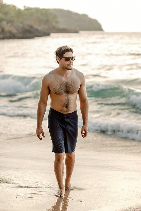 Suave man in swim shorts walks Dominican beach, gazing intently., by Cavan Images / Samantha Joy Shantz Photography