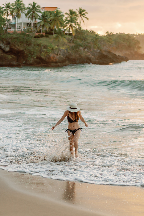 Joyful woman in black bikini and hat plays in surf at tropical sunset., by Cavan Images / Samantha Joy Shantz Photography