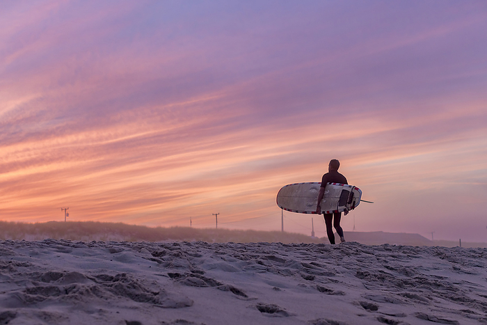 A surfer walks on the beach with his surfboard at sunset, by Cavan Images / Julia Cumes