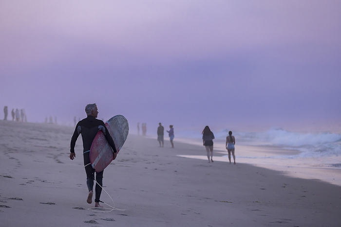 An older surfer walks on the beach with his surfboard on Cape Cod, by Cavan Images / Julia Cumes