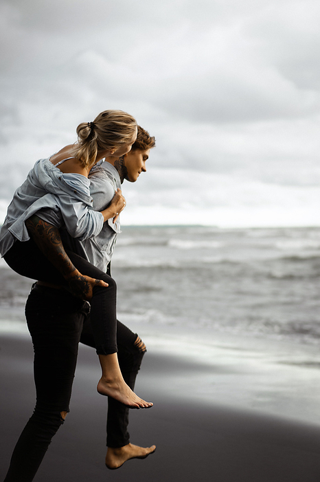 Man piggybacking woman while walking on boardwalk at beach, by Cavan Images / Yuliya Kirayonak