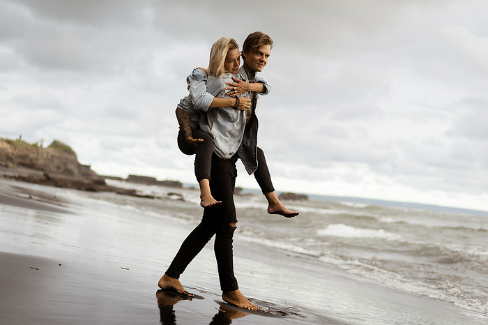 Cheerful man piggybacking woman while standing at shore, by Cavan Images / Yuliya Kirayonak