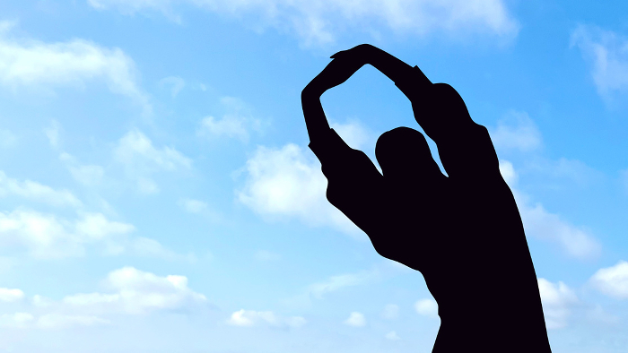Silhouette of woman with fingers of both hands crossed and stretched above her head, body at an angle, upper body blue sky background