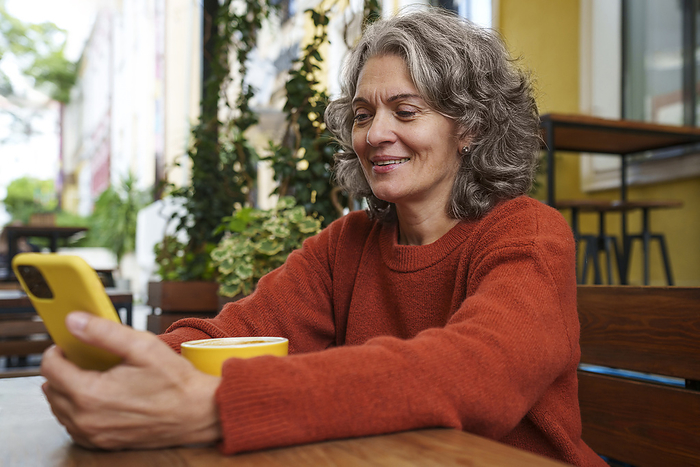 Happy mature woman using smart phone with coffee cup on table at sidewalk cafe