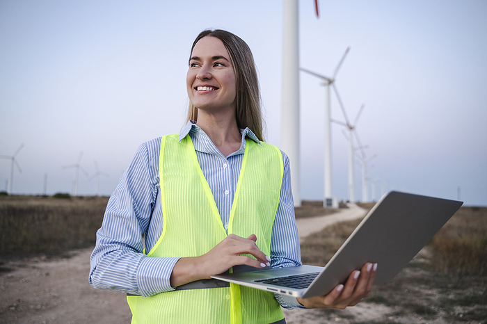 Happy engineer standing with laptop near wind turbines at field