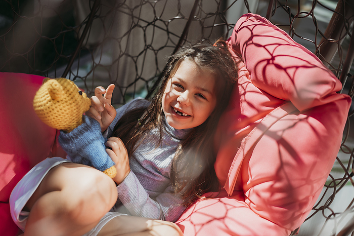 Smiling girl with stuffed toy lying on porch swing