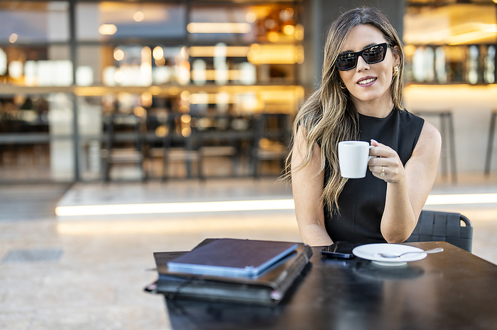 Smiling businesswoman holding coffee cup and sitting at sidewalk cafe