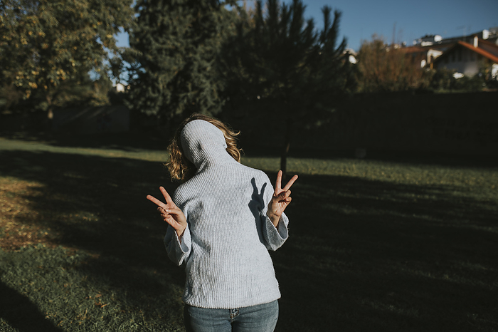 Playful woman covering face with hooded sweater and gesturing peace sign at autumn park