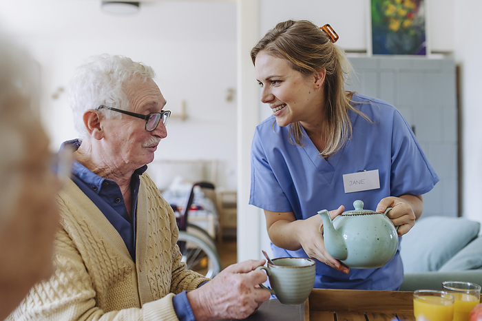Happy healthcare worker holding teapot and talking to senior man at table