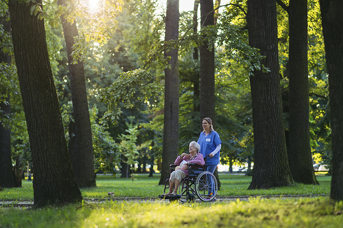 Smiling healthcare worker walking with senior woman sitting in wheelchair near trees at park