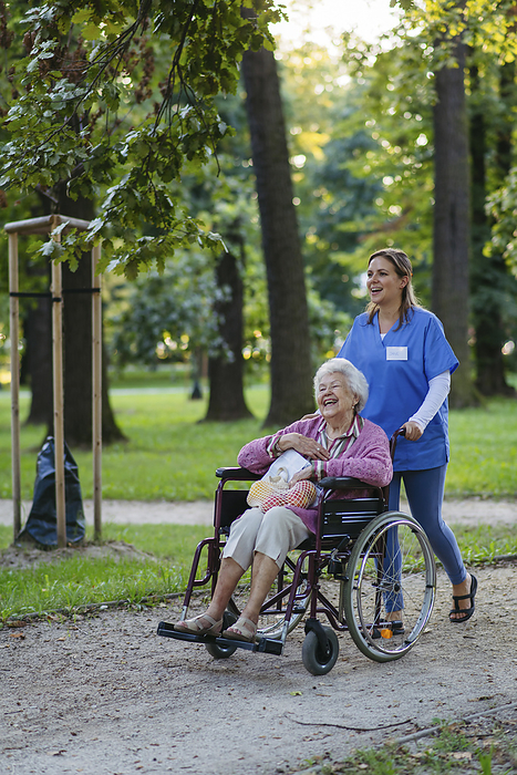 Happy healthcare worker walking with senior woman sitting in wheelchair at park