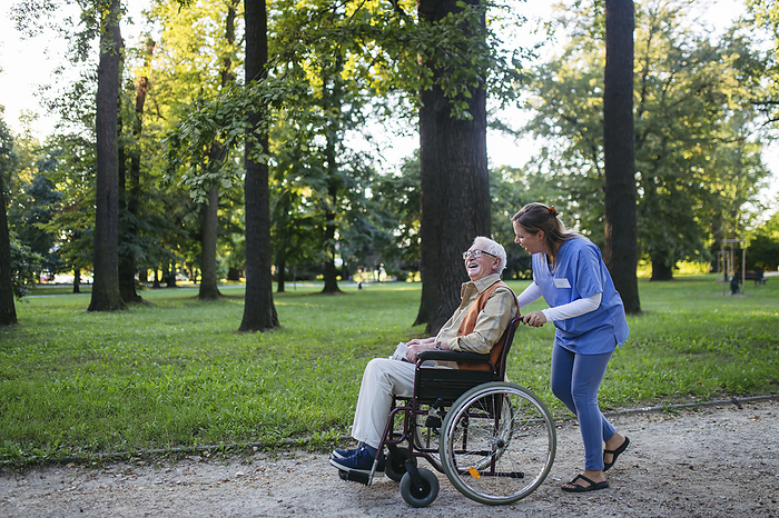 Happy healthcare worker walking with senior man sitting in wheelchair near trees at park