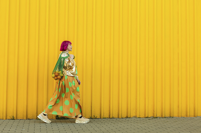 Senior woman carrying fruits in mesh bag and walking near yellow wall