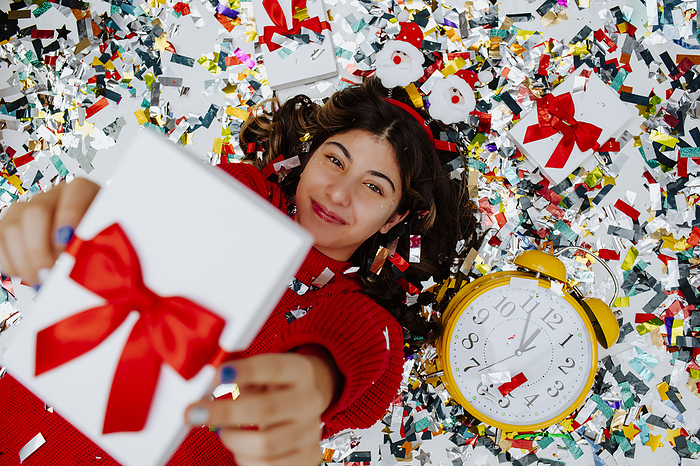 Smiling girl holding gift box and lying on confetti near clock