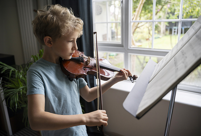 Elementary boy practicing to play violin near window at home