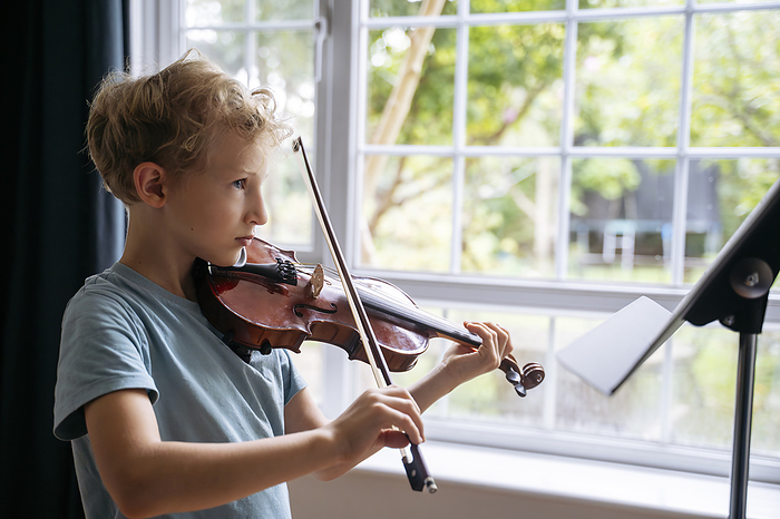 Boy practicing violin with sheet music near window at home