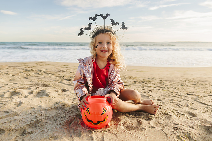 Smiling girl sitting with Halloween pumpkin toy on sand at beach