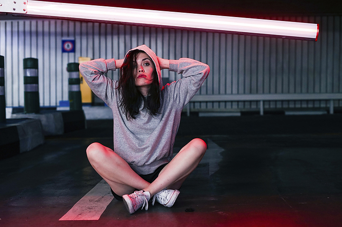 Woman wearing hooded shirt sitting on ground under neon lighting