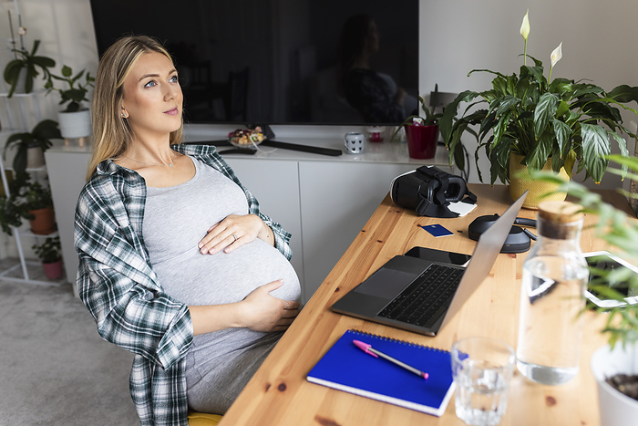 Contemplative pregnant freelancer sitting with laptop at desk in home office