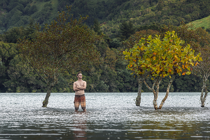 Shirtless man standing in lake with arms crossed near trees